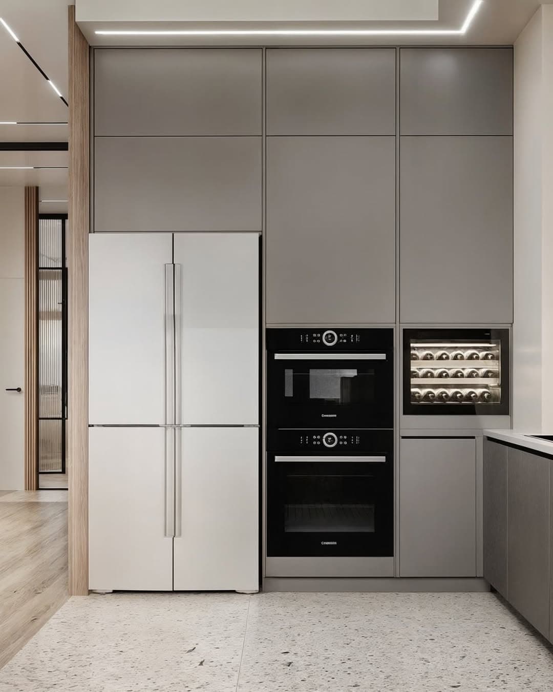 Modern minimalist kitchen with floor-to-ceiling taupe cabinet panels, white French-door refrigerator, built-in black ovens stacked vertically, and integrated wine cooler with visible bottles. Terrazzo-style flooring with flecked pattern completes the contemporary design.