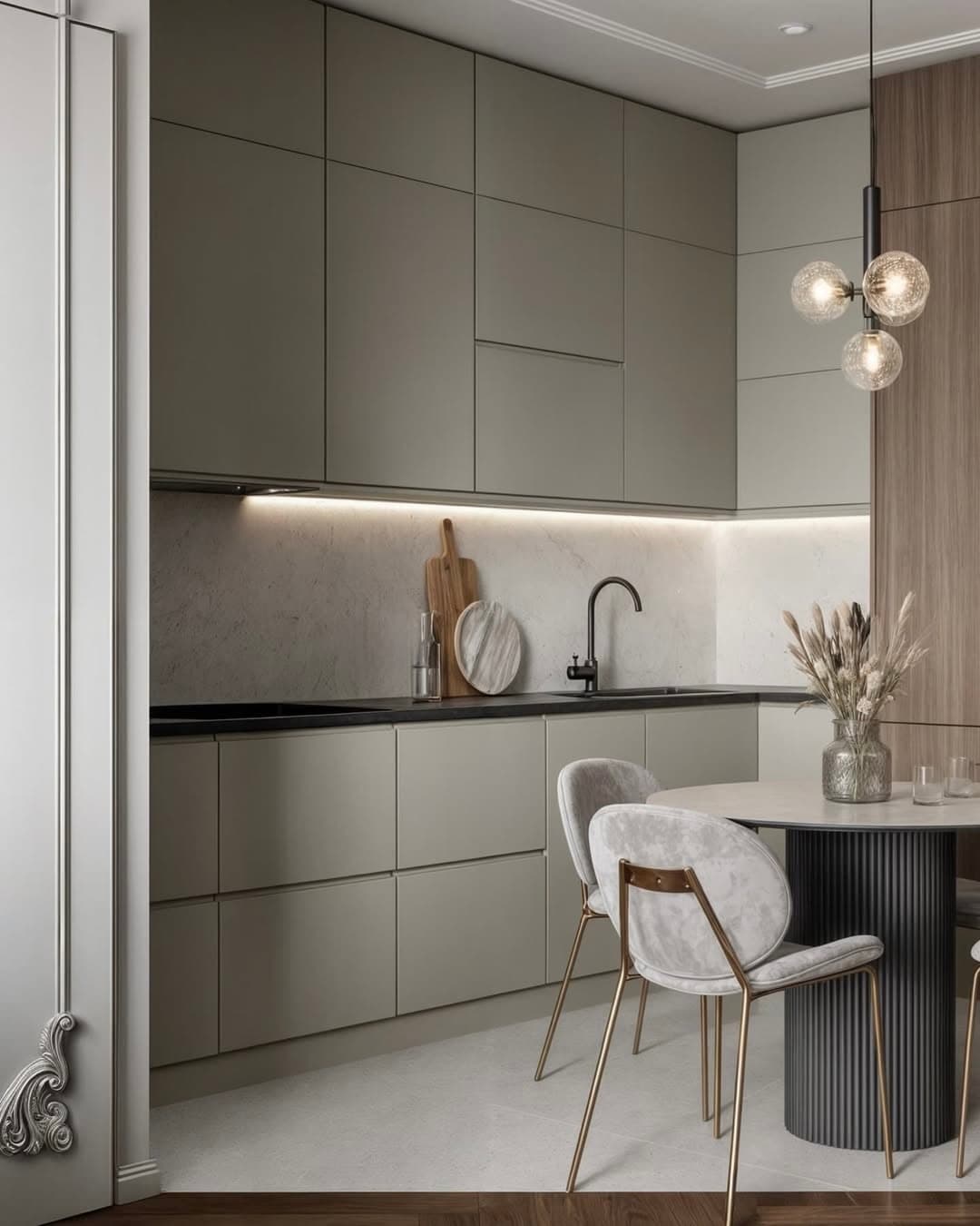 Elegant kitchen with sage green flat-panel cabinets, black countertops, white marble backsplash with LED lighting, modern globe pendant light, and round breakfast table with gray velvet chairs.